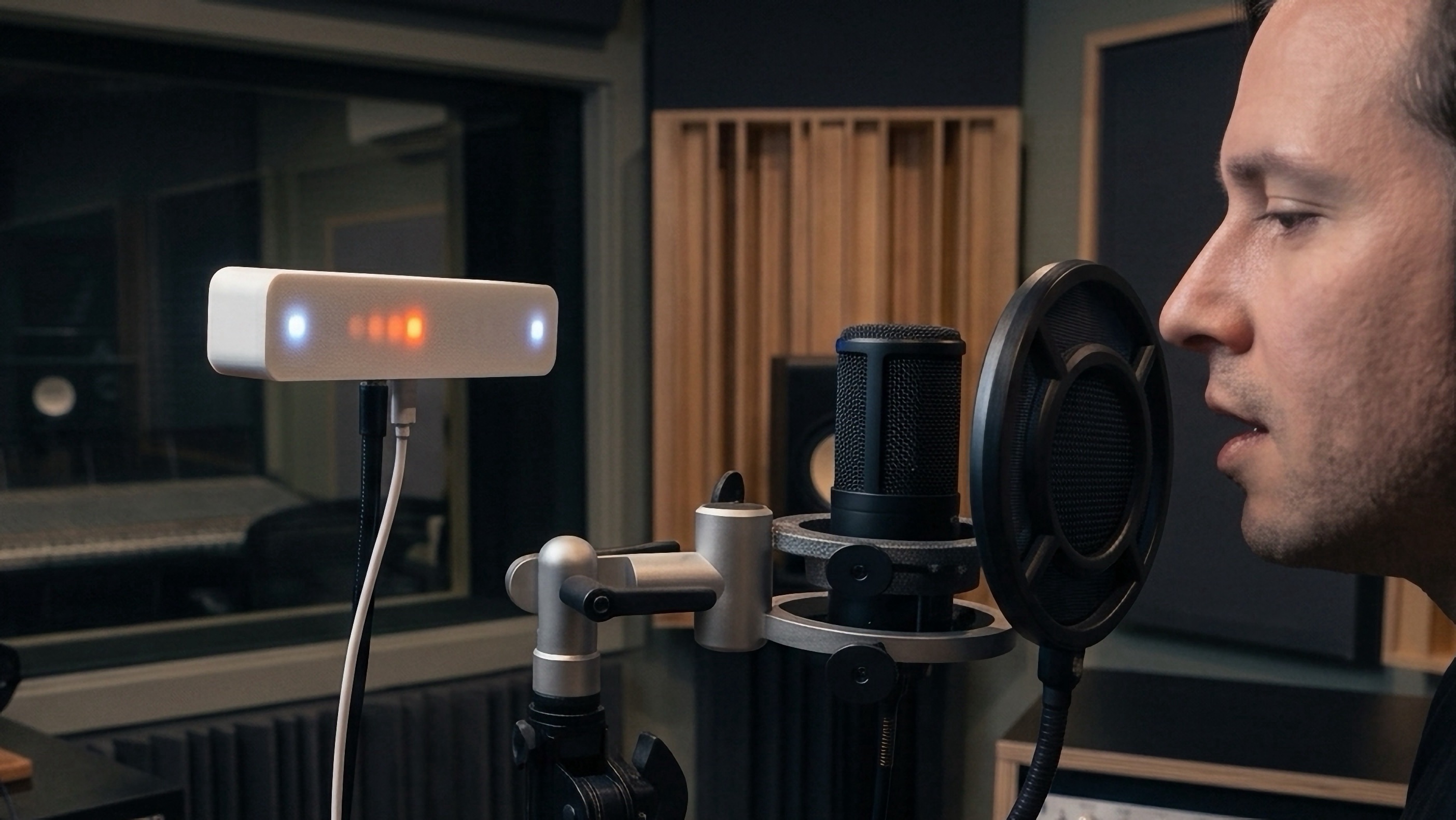 A Vistanome on a mic stand in a recording studio, its LED strip lit, as a vocalist sings into a condenser mic